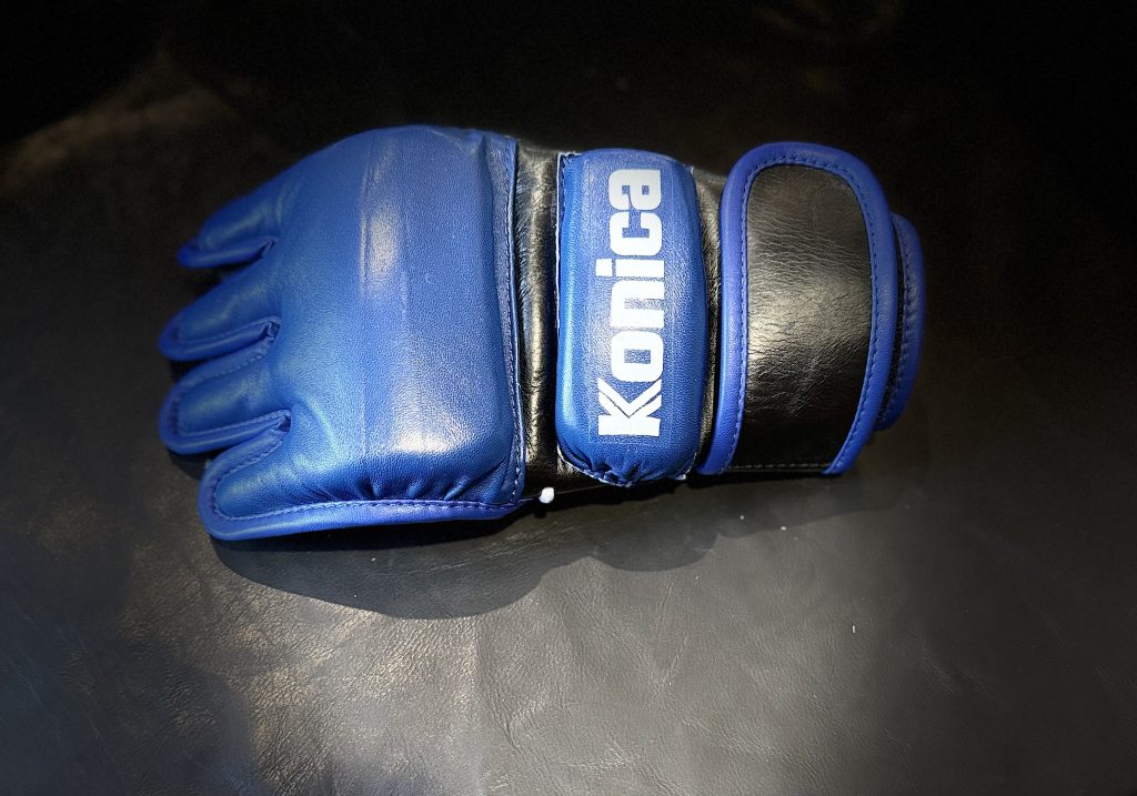Replica blue Konica MMA gloves worn by Dwayne Johnson as Mark Kerr in The Smashing Machine, costume by Heidi Bivens.